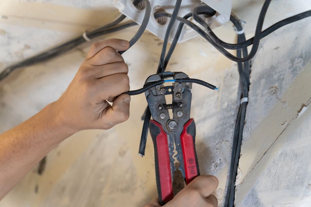 Person stripping insulation from a black electrical wire with red and gray wire strippers.