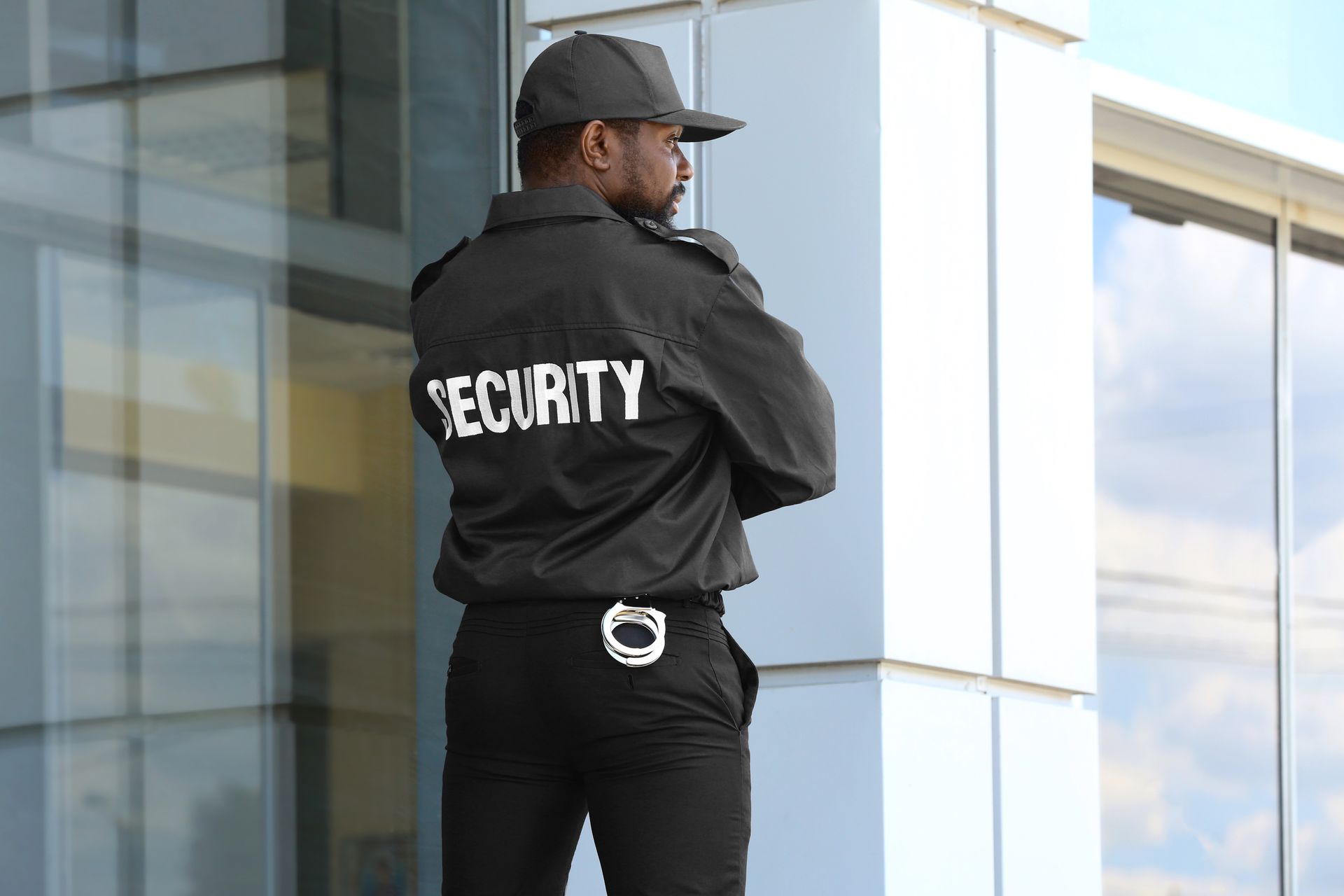 Security guard standing outside a modern glass building, wearing a black jacket and cap.