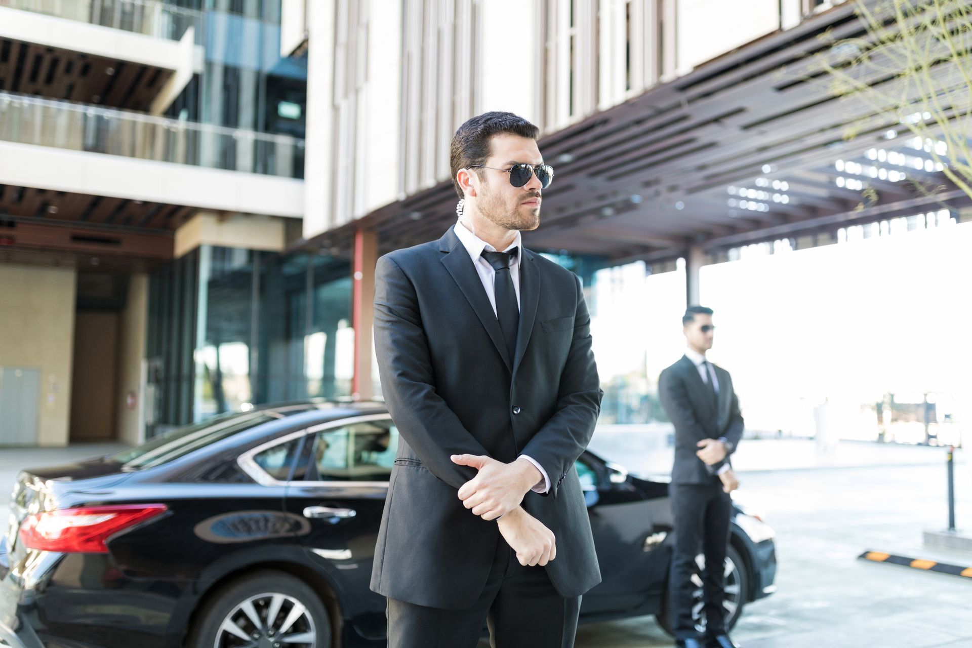 Two suited security guards stand beside a black car outside a modern building.