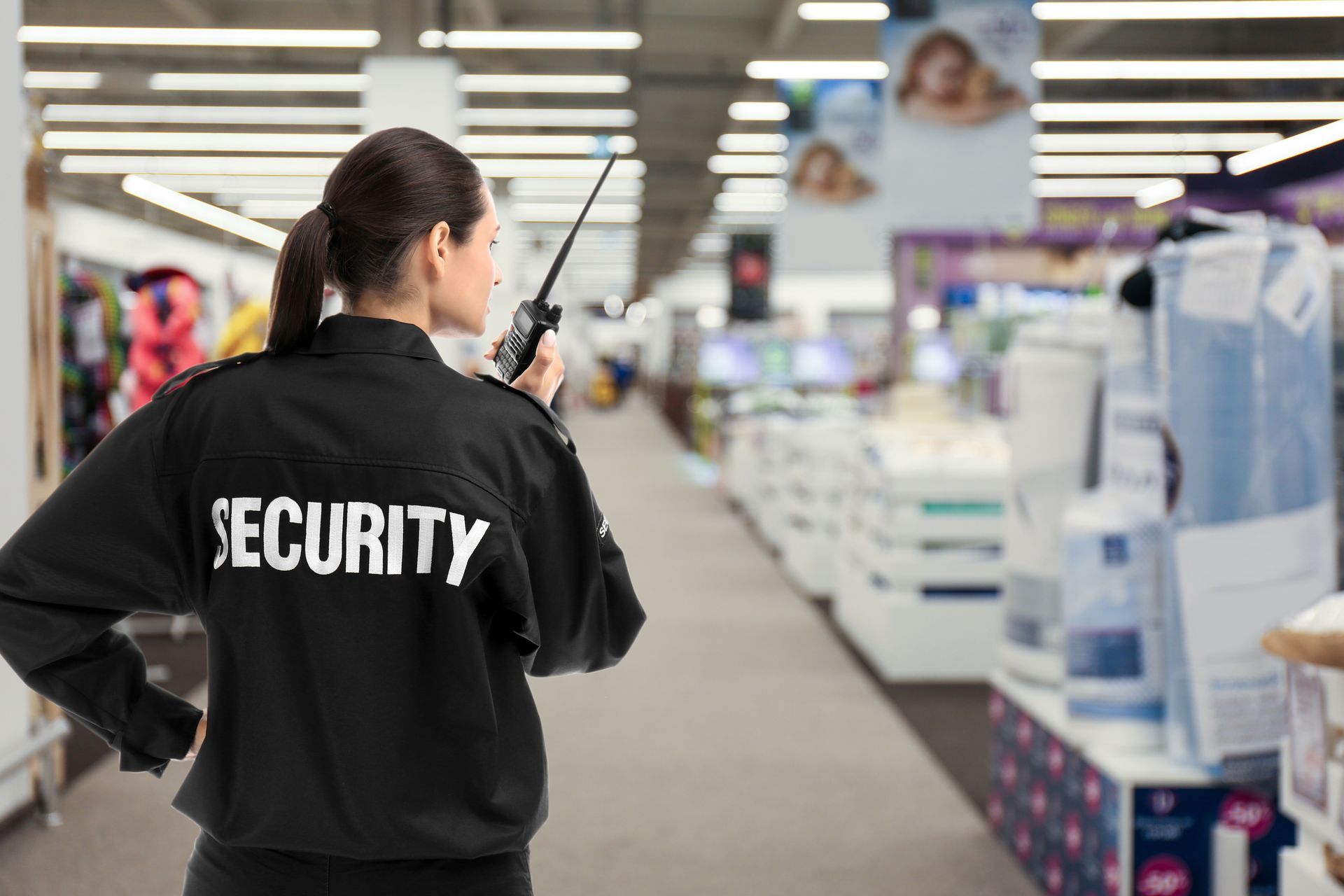 Security guard in black jacket with radio, walking through a store aisle.