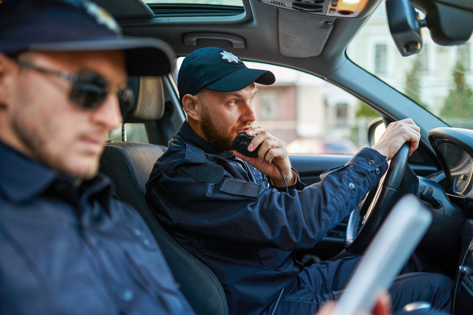 Two police officers in a patrol car, one driving and one looking at a device.