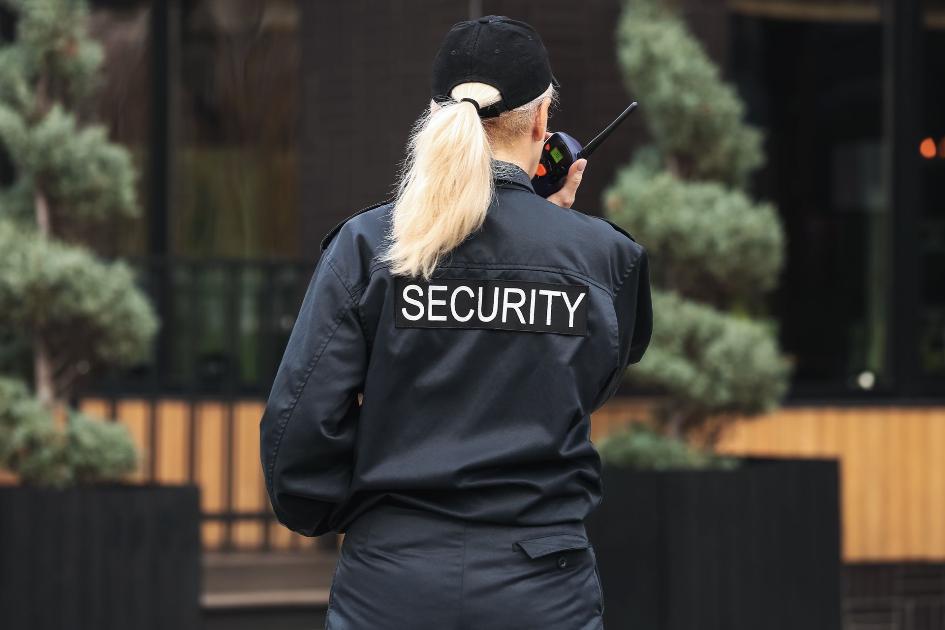 Security guard in black jacket and cap speaking into a walkie-talkie outdoors