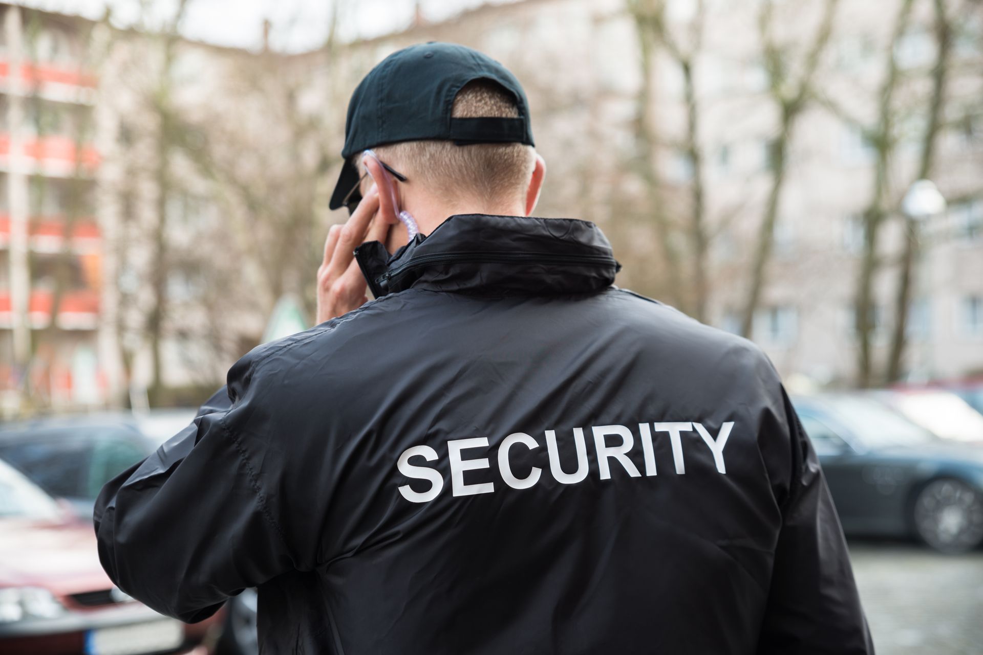 Security guard in black jacket and cap talking on a phone outdoors, with cars blurred in the background
