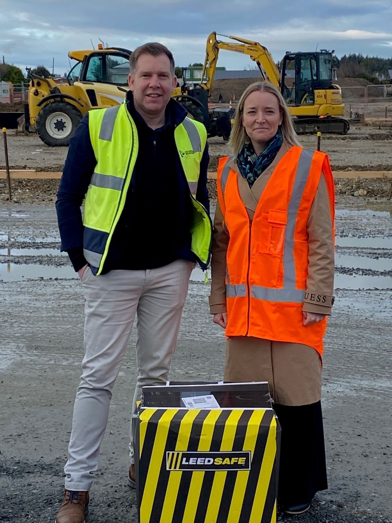 Two people in safety vests at a construction site stand beside a black and yellow labeled container.