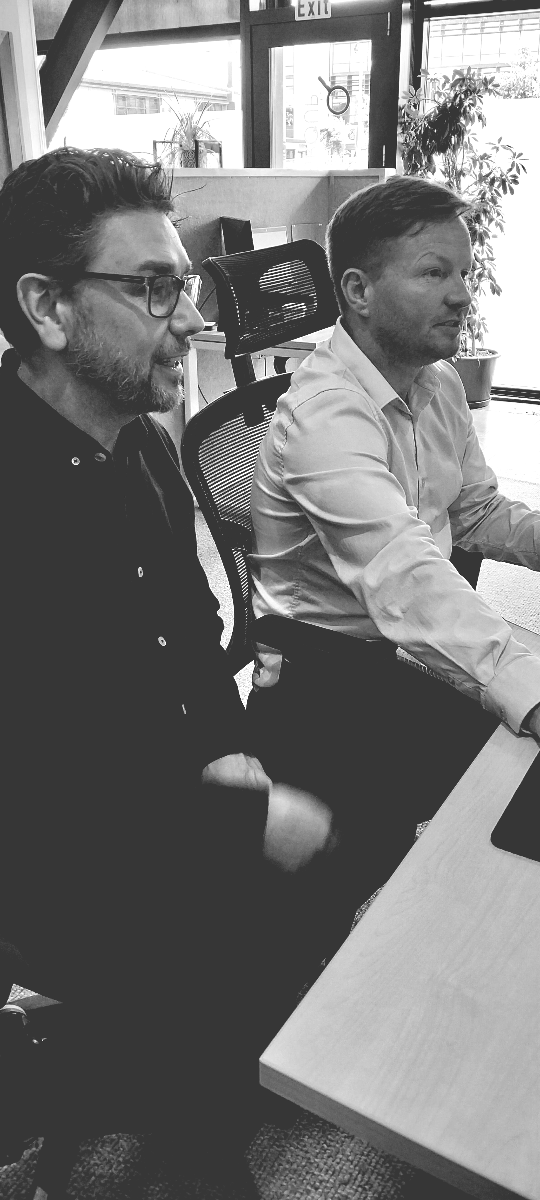 Two men seated at a table in an office setting, both looking to the right. One wears glasses and a dark shirt.