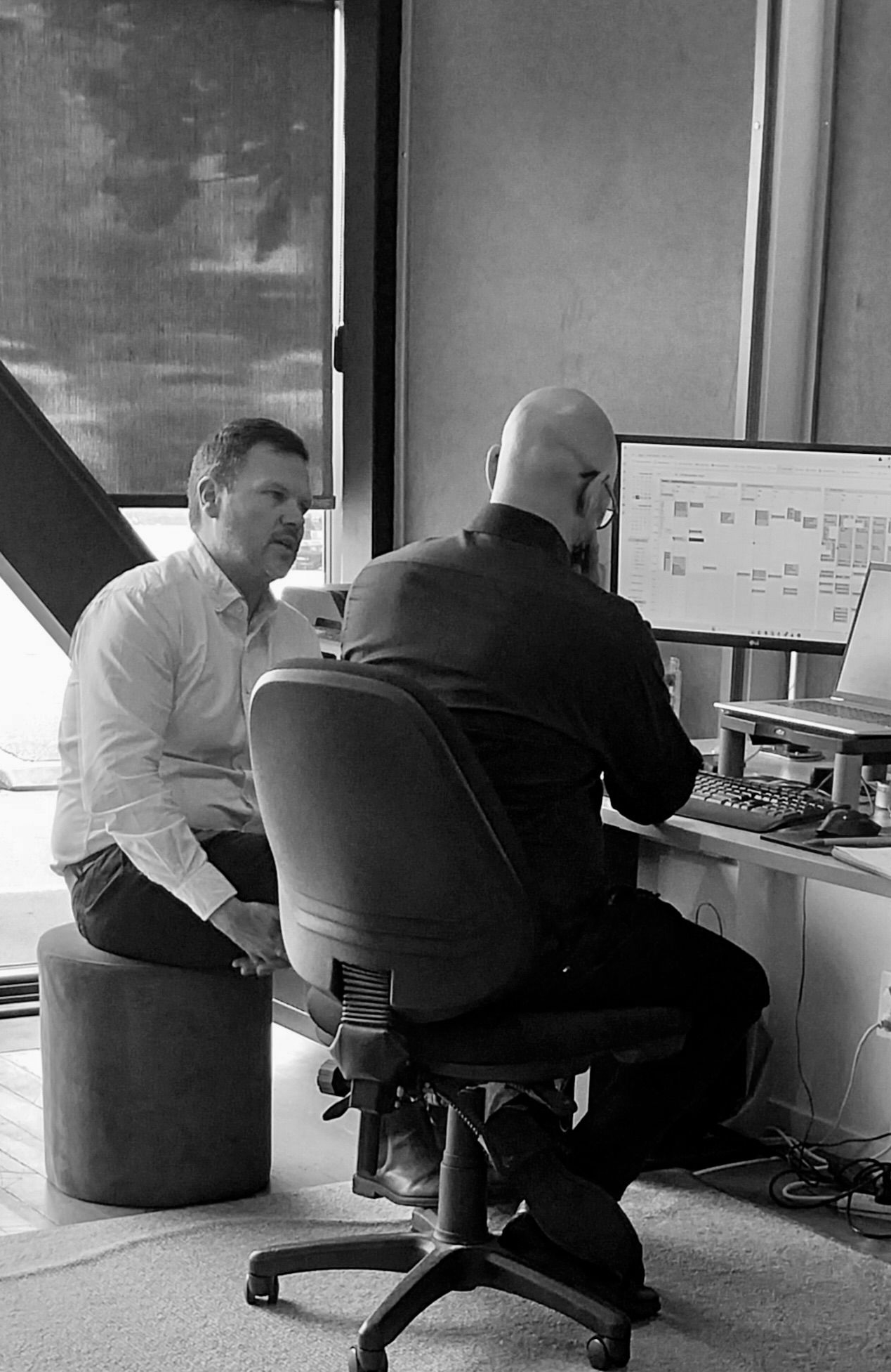 Two men working together at a desk. One sits in a chair, looking at a computer screen. The other sits on a stool, leaning in.