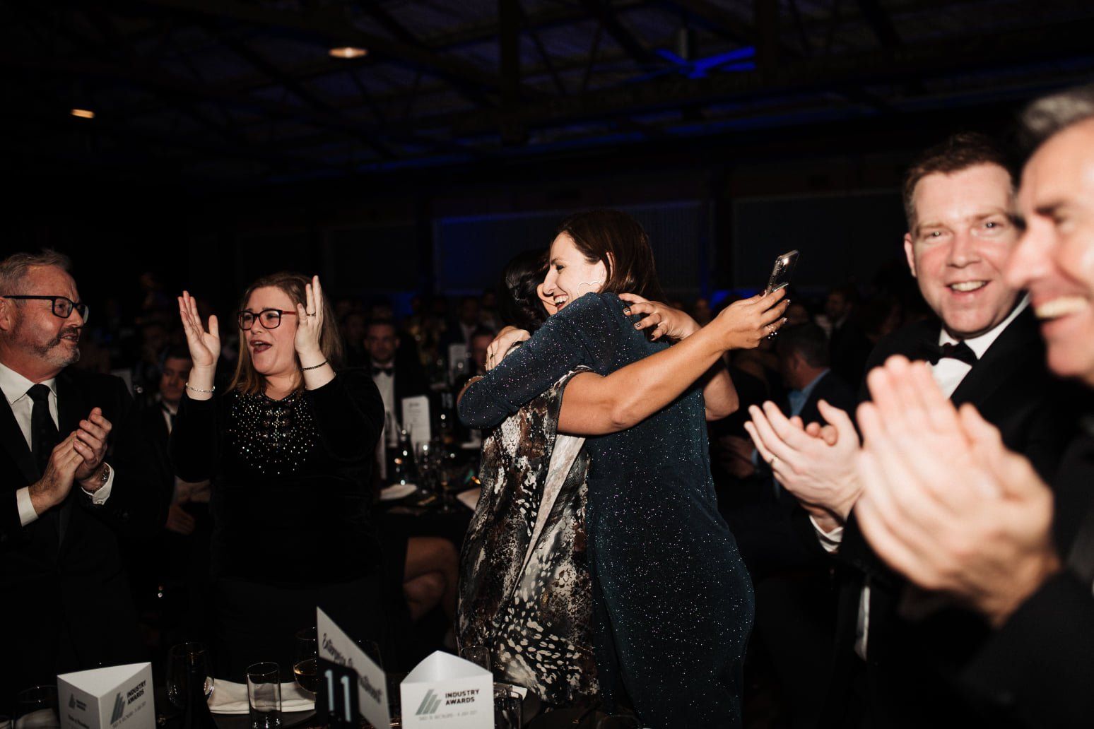 People hug and clap at an awards ceremony. A woman in a dark dress holds a trophy.