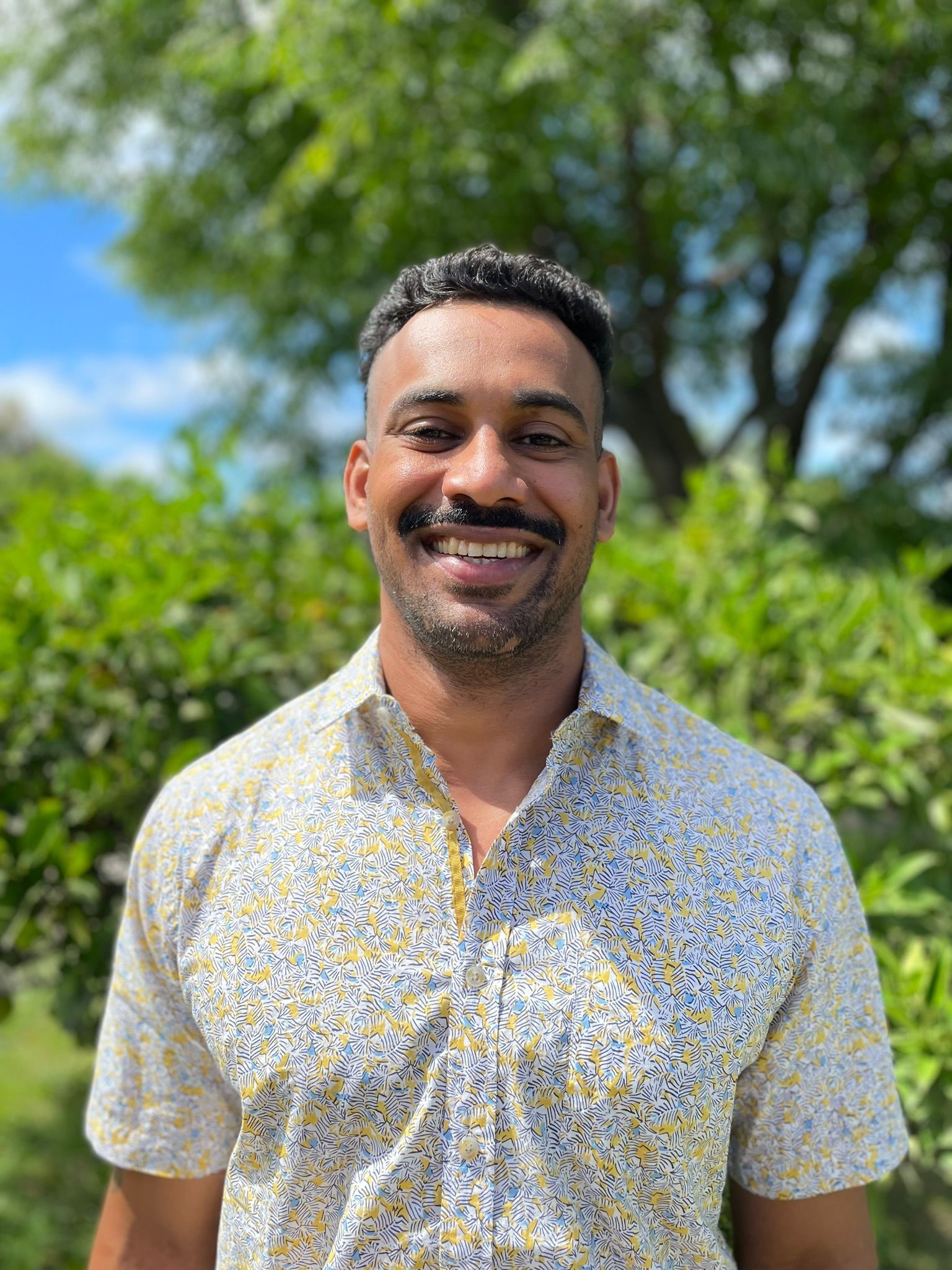 Man smiling, wearing patterned shirt, standing in front of green foliage.