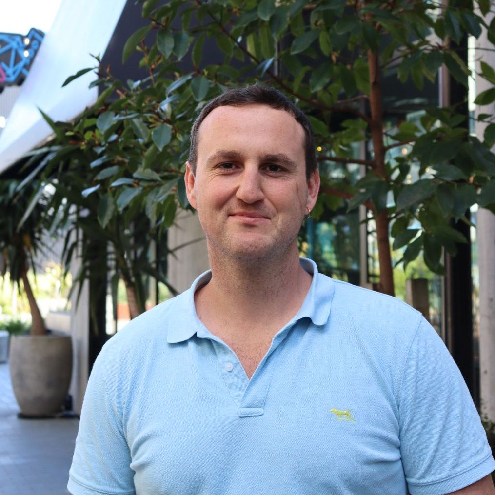 Man in a light blue polo shirt smiles outside. Trees and a modern building are in the background.