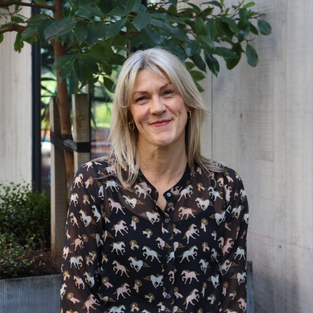 Woman with blonde hair wearing a horse-print shirt smiles outdoors by a building and plants.