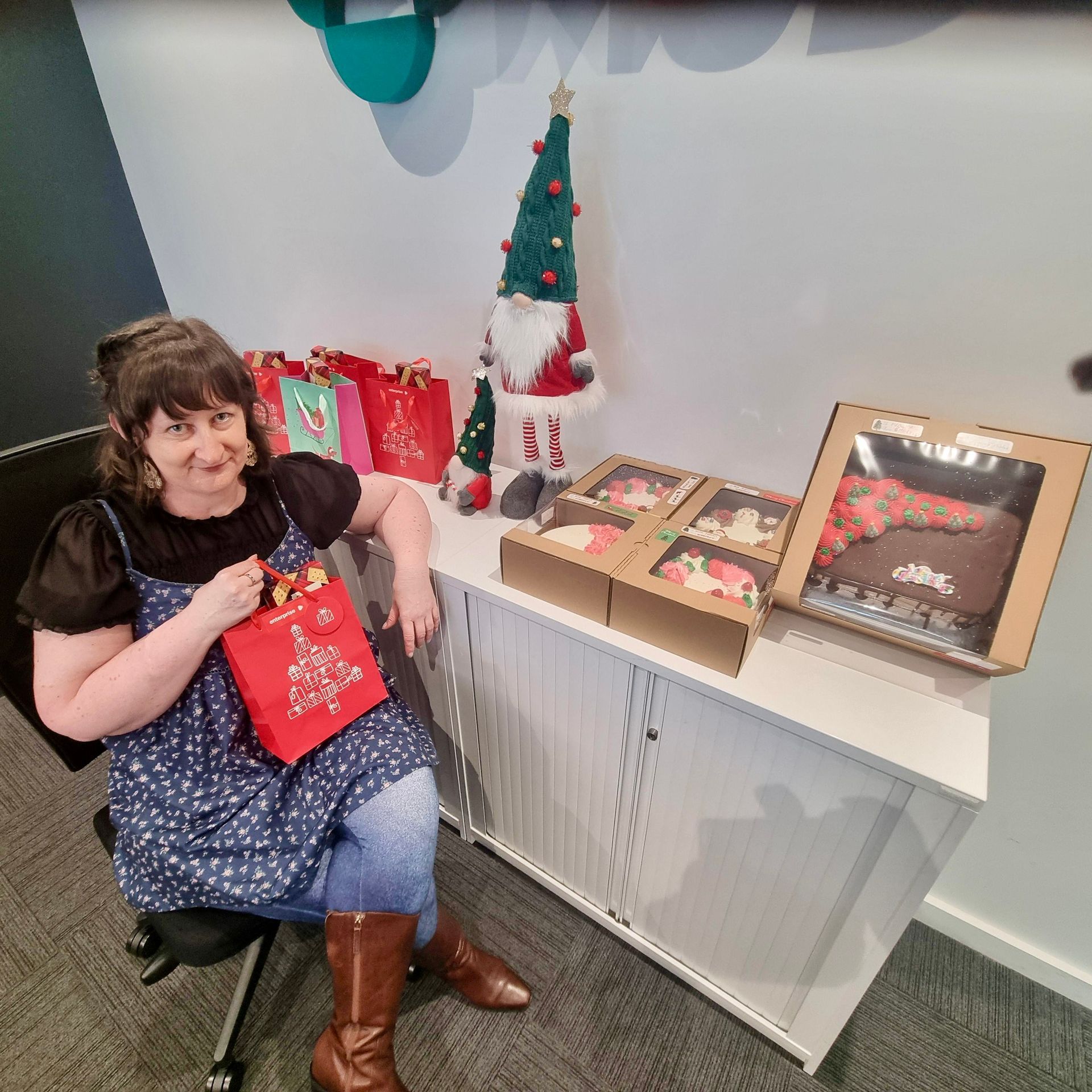 Woman in floral dress and boots, seated next to desk with Christmas gifts and decorations.