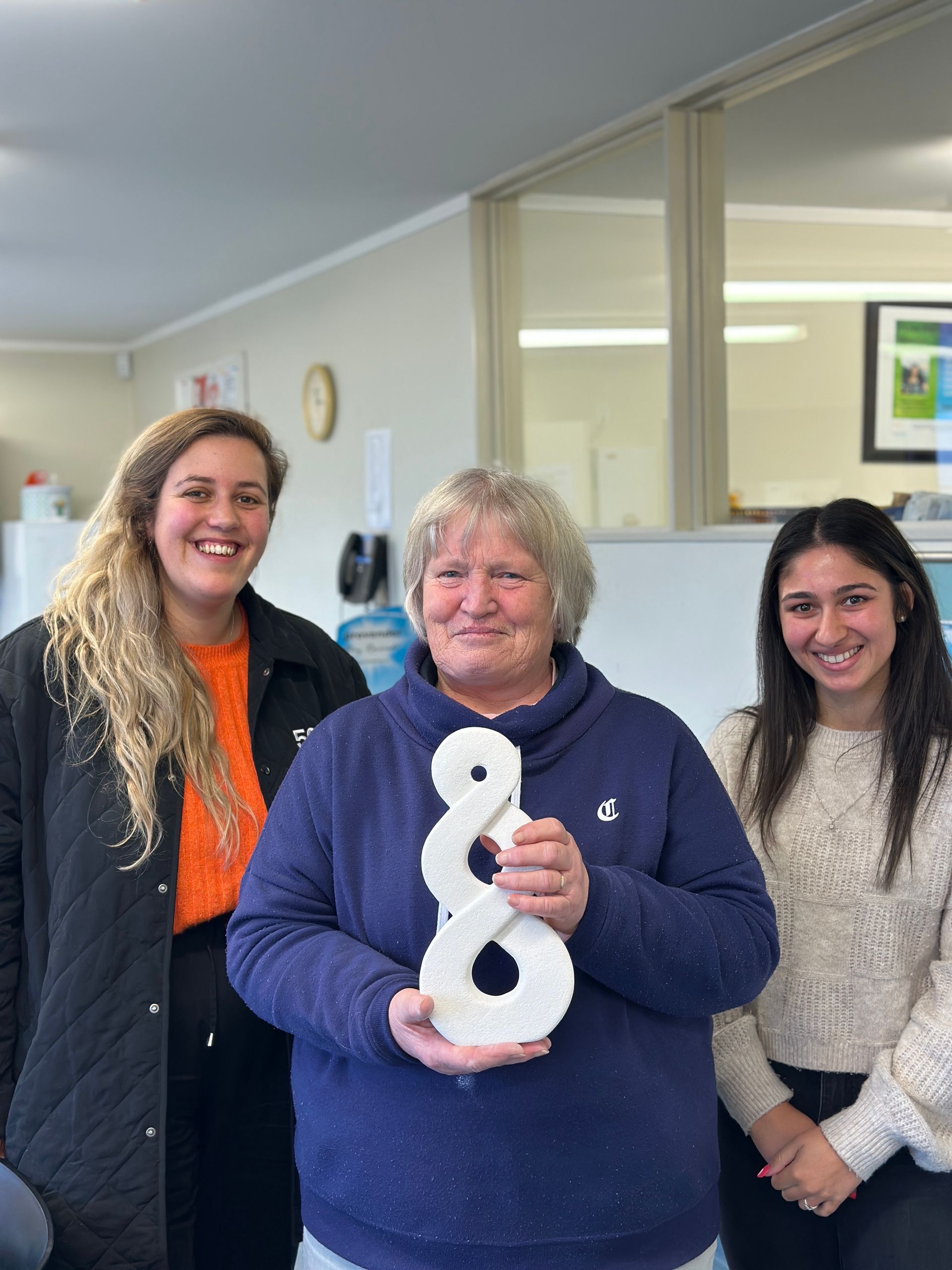 Three people inside; one holds a white, figure-eight-shaped sculpture. All smiling, light interior with neutral tones.