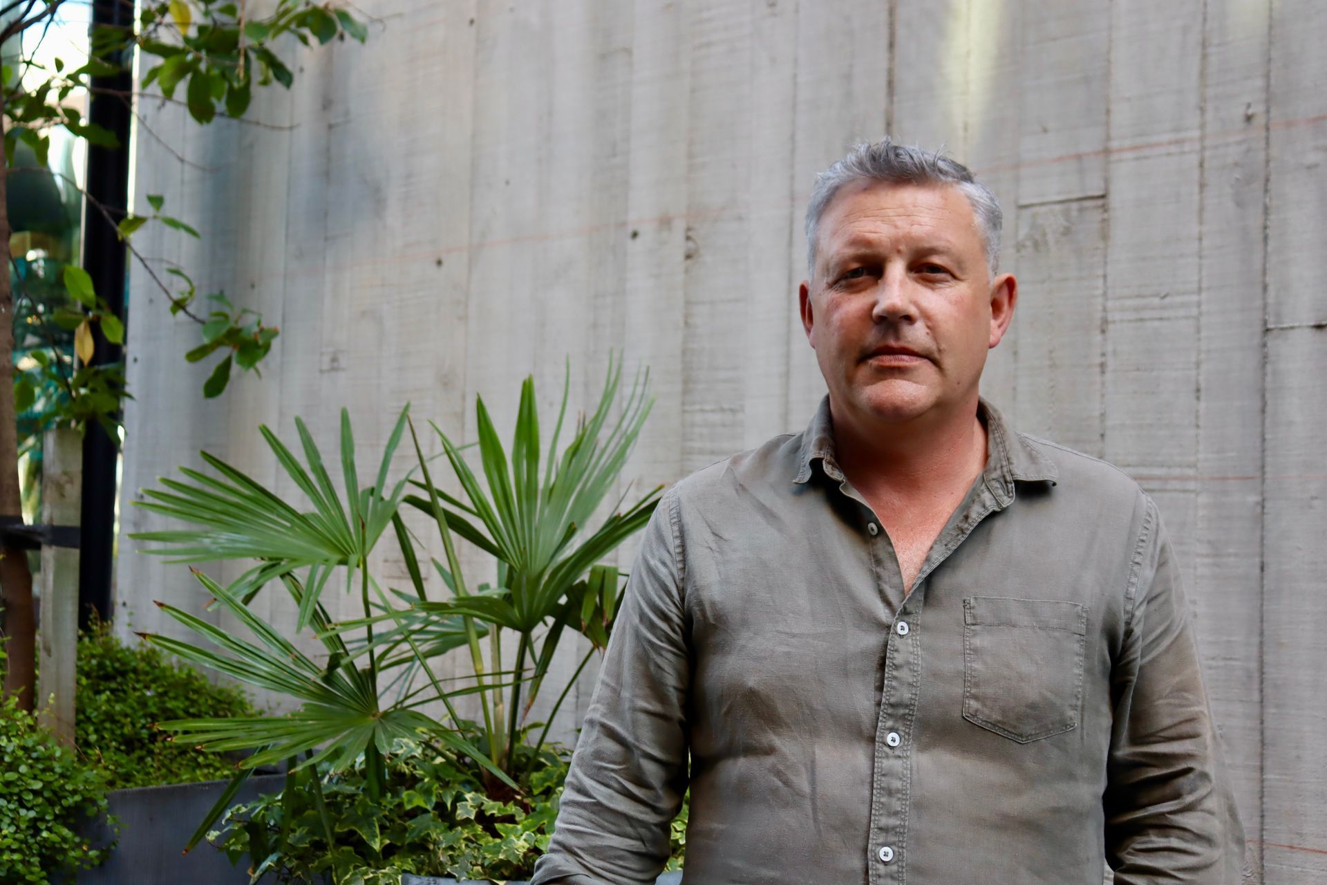 Man standing outdoors in front of a concrete wall and green plants, wearing a gray shirt.