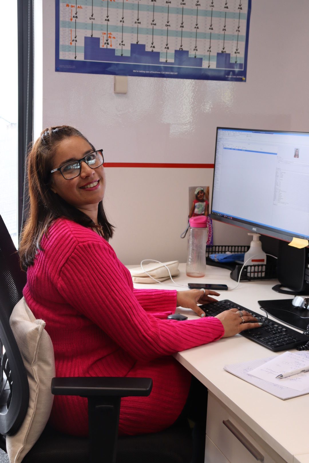 Woman in pink sweater smiling while typing at a desk with a computer.
