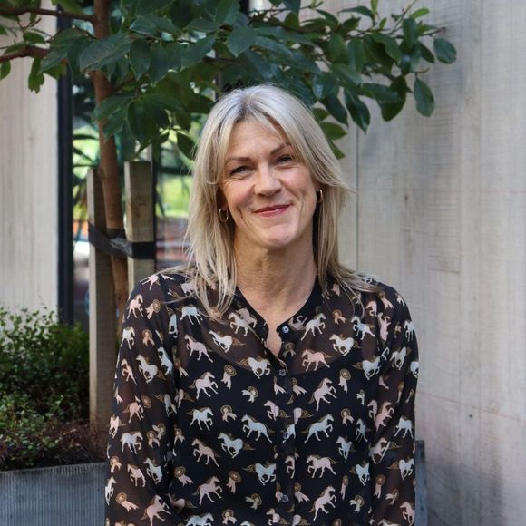 Woman with blonde hair, smiling in front of a tree, wearing a horse-print blouse, outdoors.