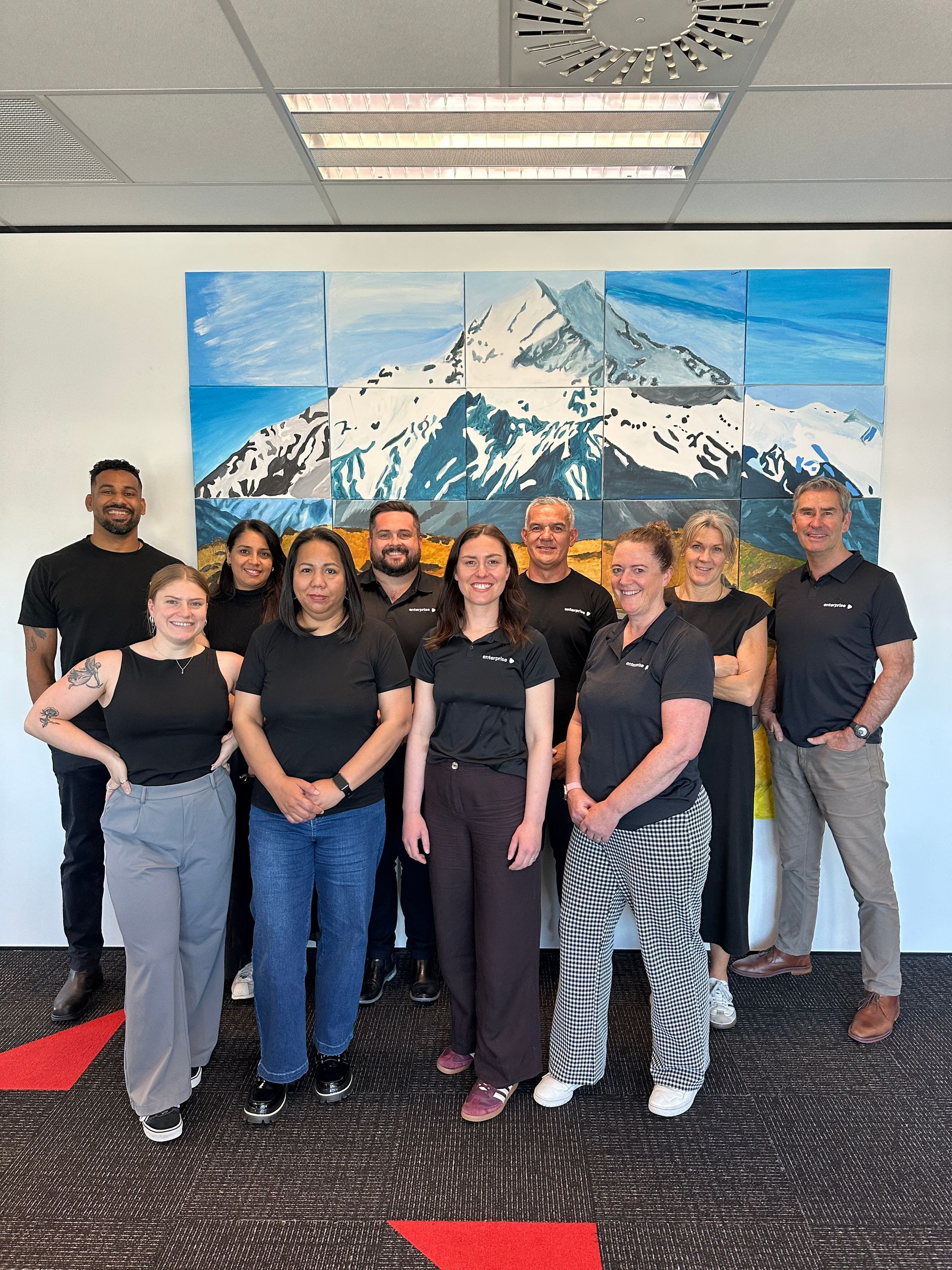 Group of people posing in front of a mountain painting. All are wearing black shirts. Office setting.