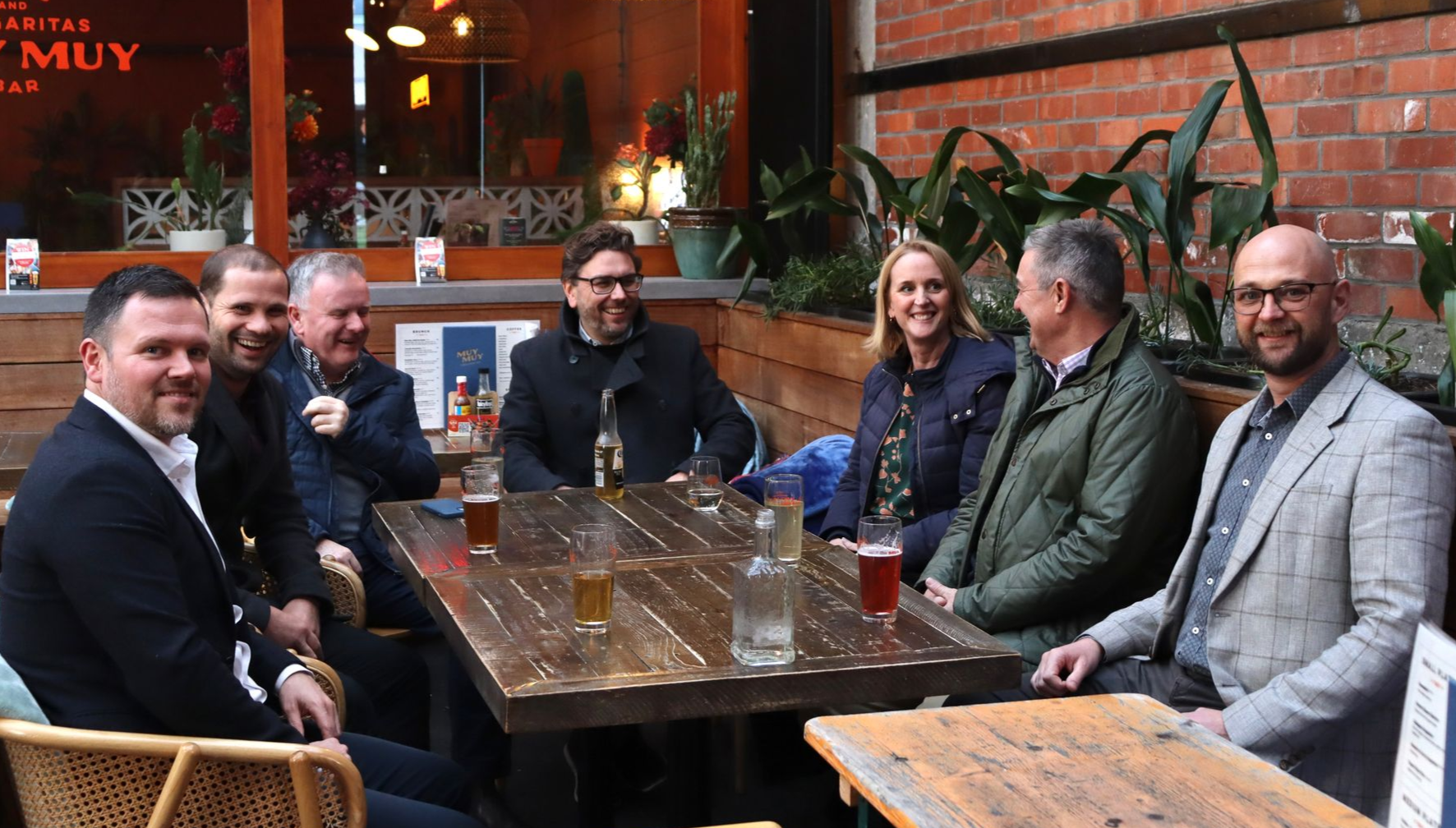 Group of people seated at an outdoor table, smiling and chatting. Beers and water on the table, brick wall backdrop.