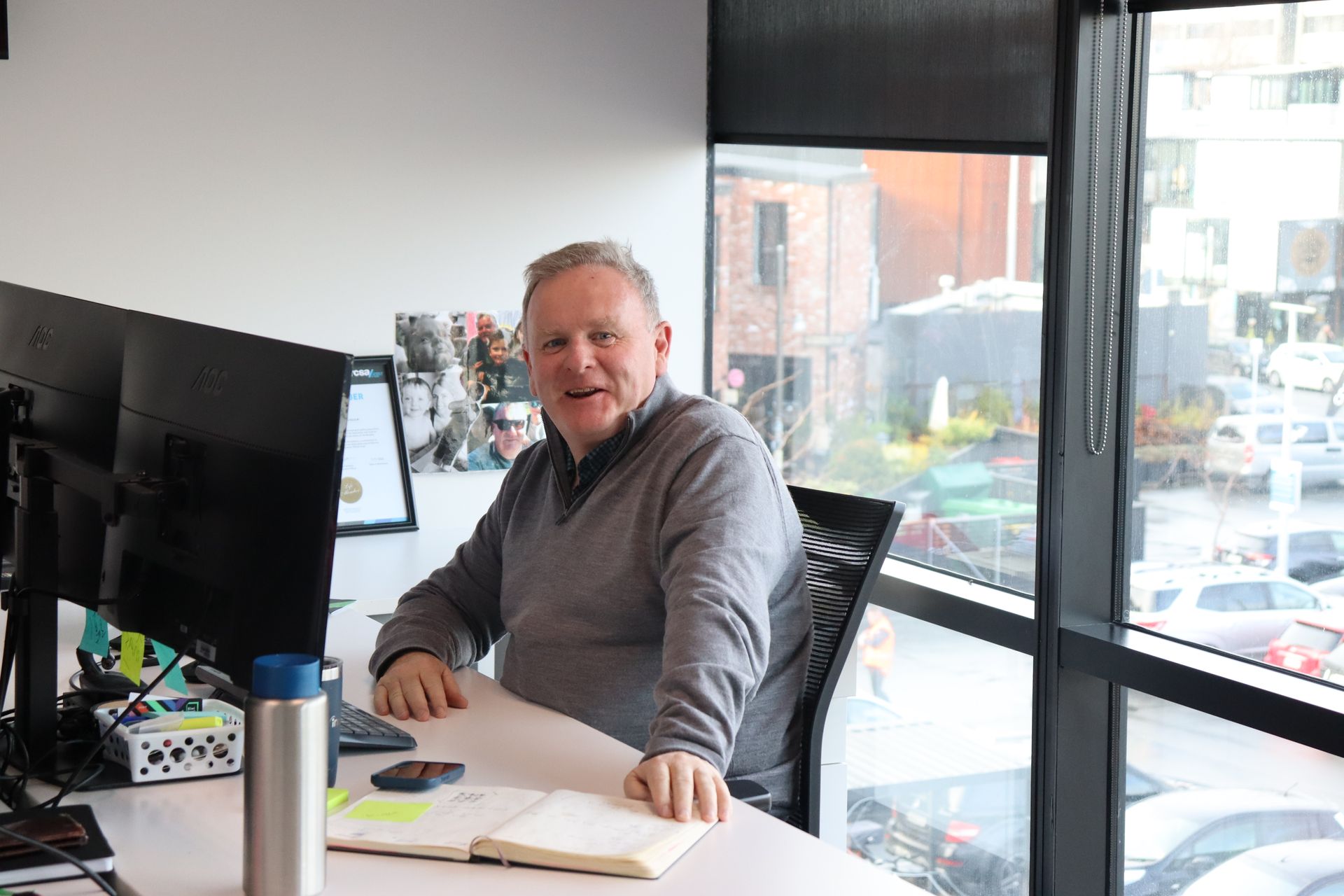 Man in gray sweater smiles, sits at desk with dual monitors, by a window.