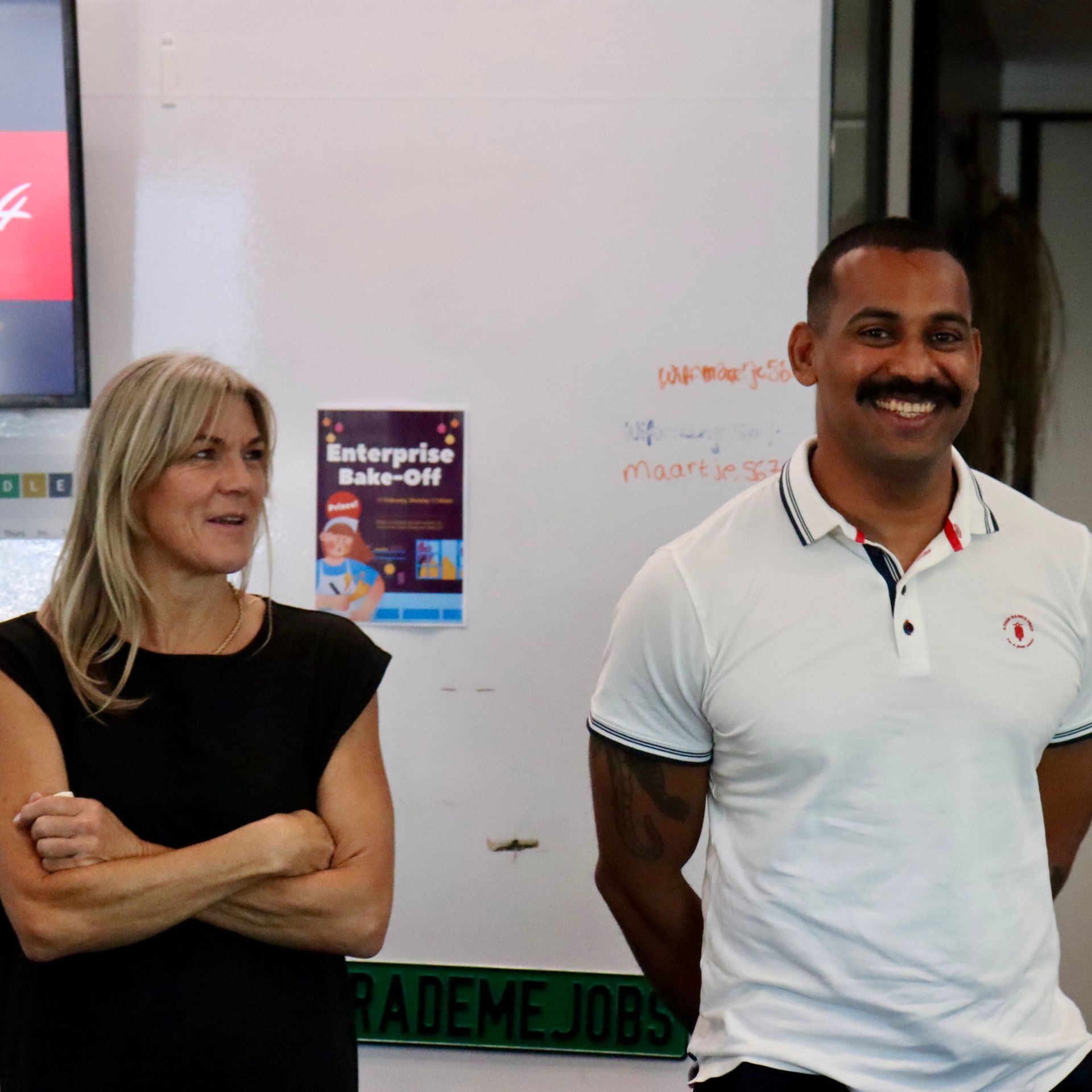 Woman and man standing near a whiteboard. The man is smiling and wearing a white polo shirt with tattoos.