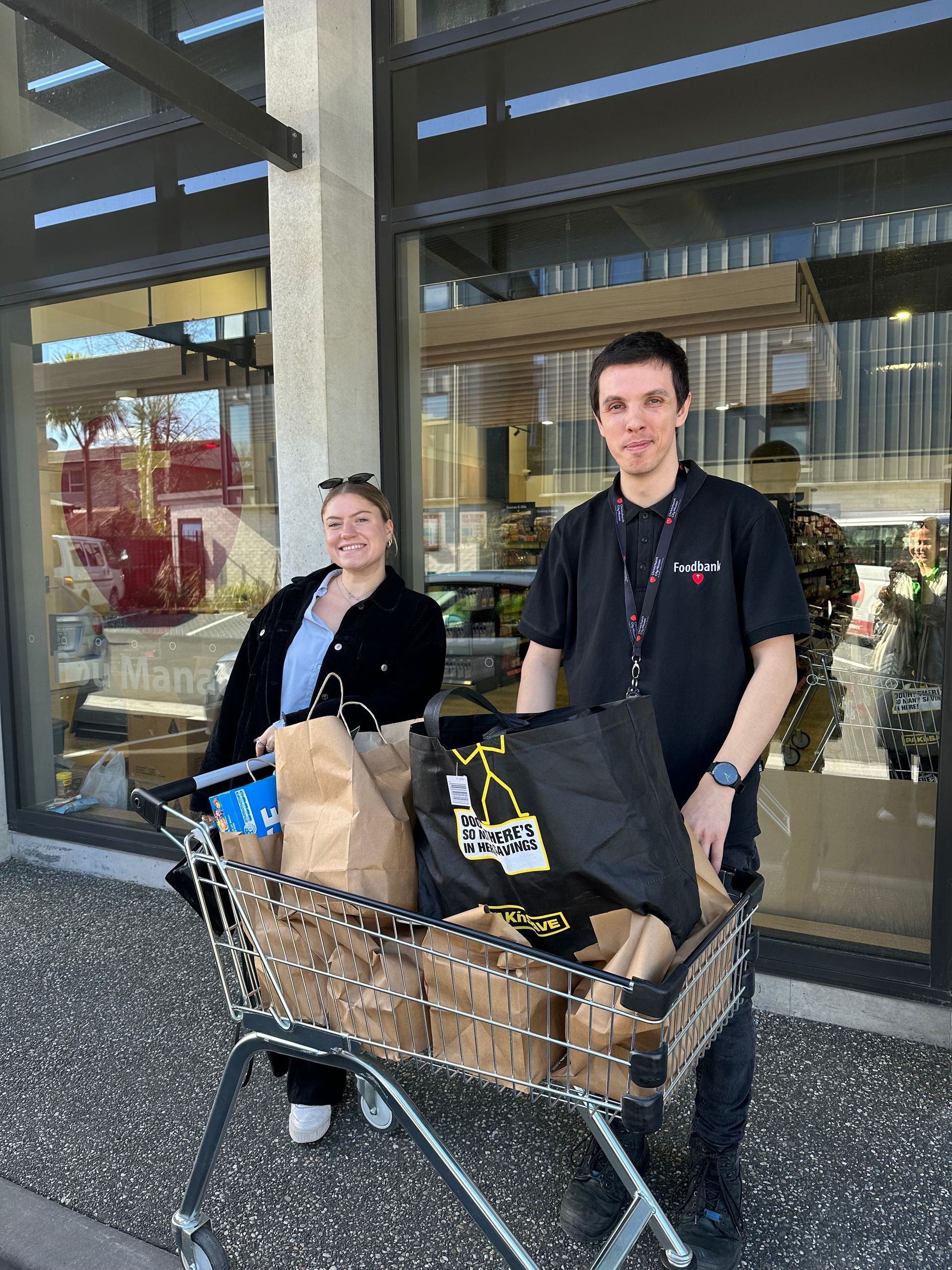 Two people with bags of groceries in a shopping cart outside a building. One person is smiling.