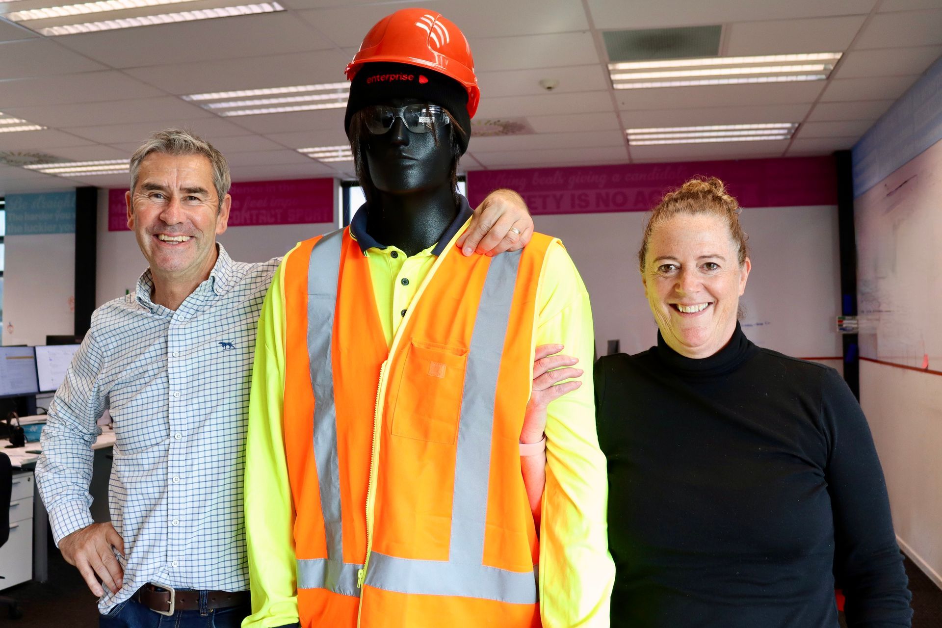 Two people flanking a mannequin in work safety gear in an office. One has arm around the mannequin.