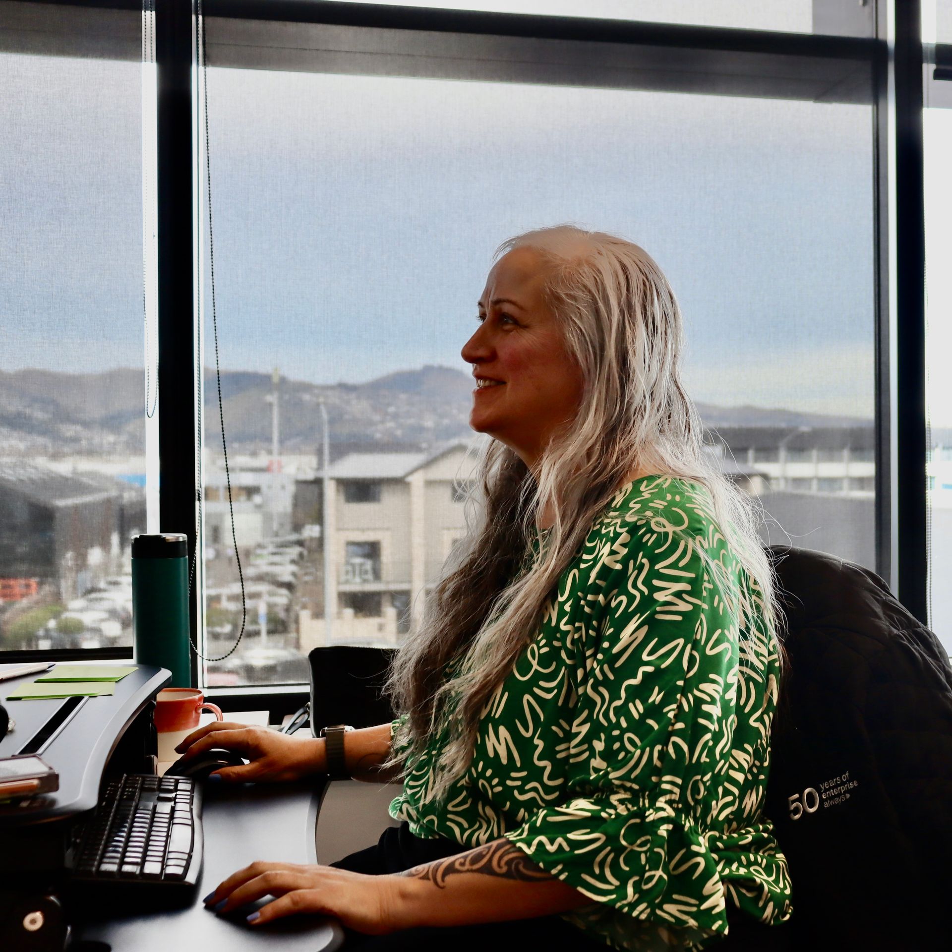 Woman with long gray hair smiles, working at a computer by a window overlooking a cityscape.