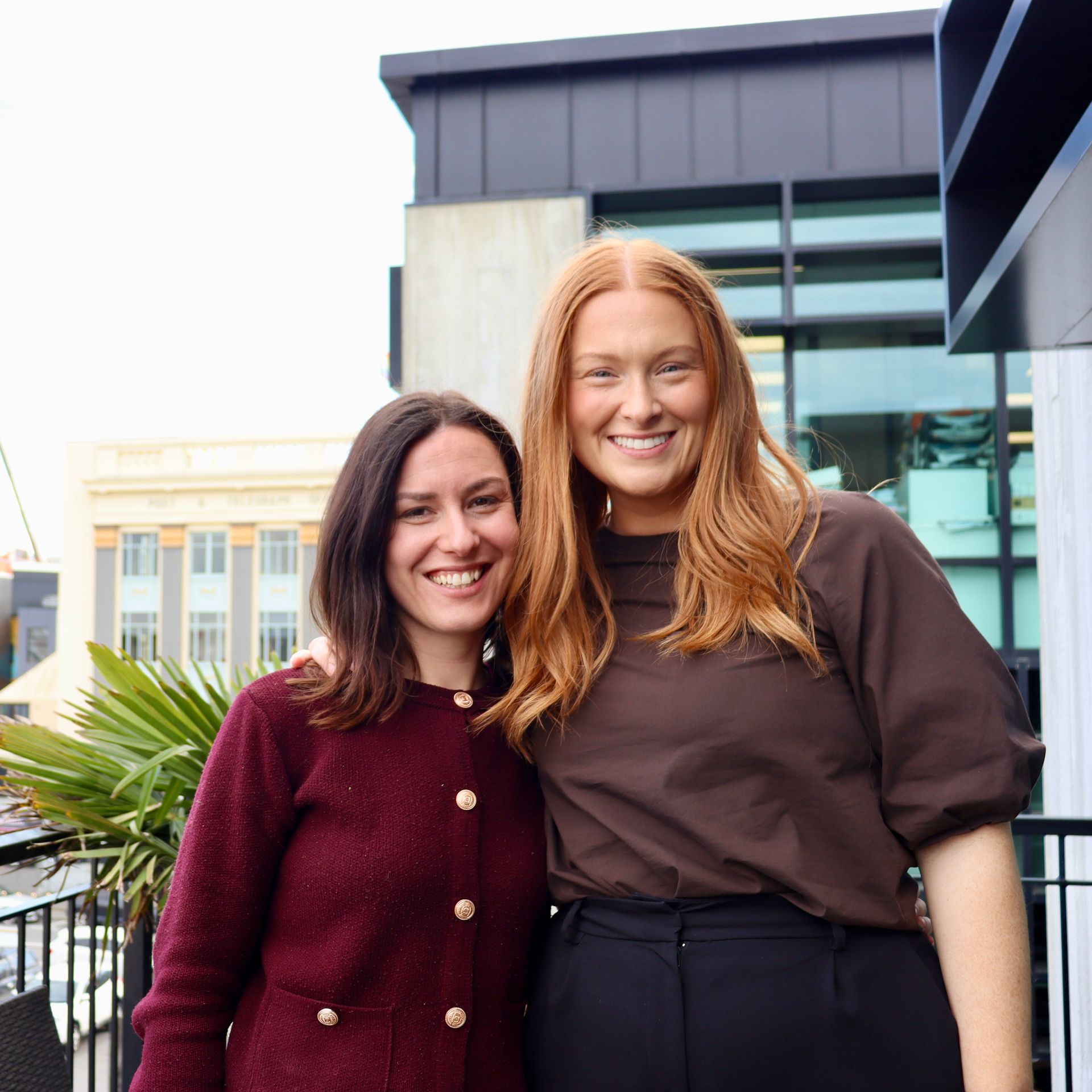 Two women smiling, arms around each other, on a balcony. One with red hair in brown top, other in a burgundy sweater.