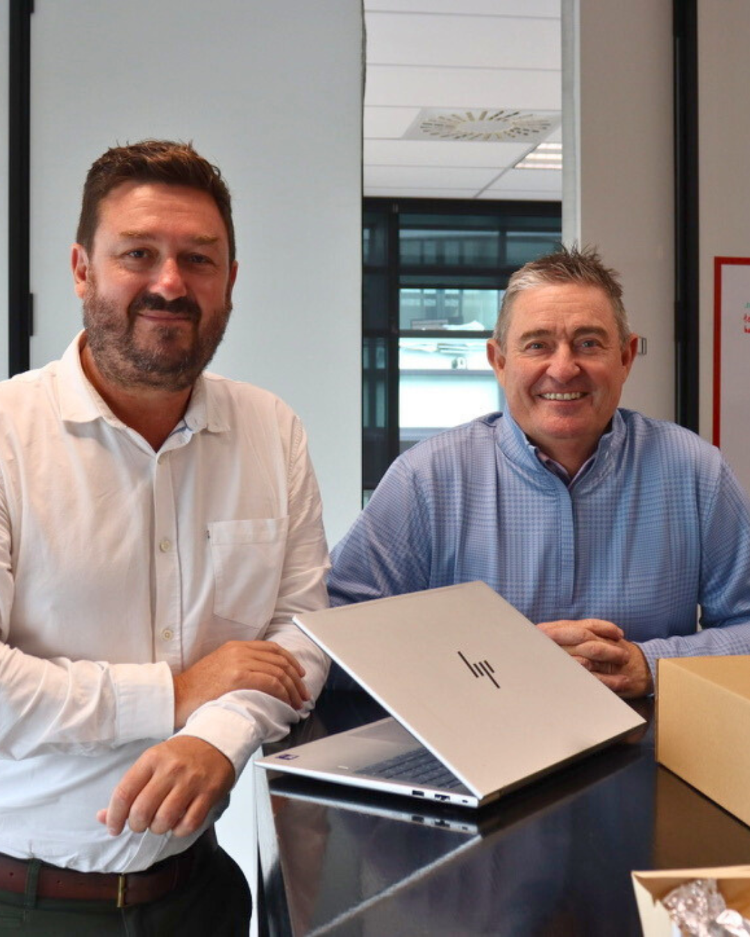 Two men beside a laptop; one wears a white shirt, the other a blue sweater, in an office.