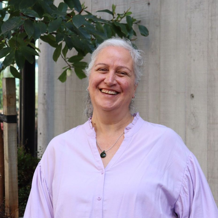 Woman with grey hair smiles, wearing a lavender shirt, standing near a wall and tree.