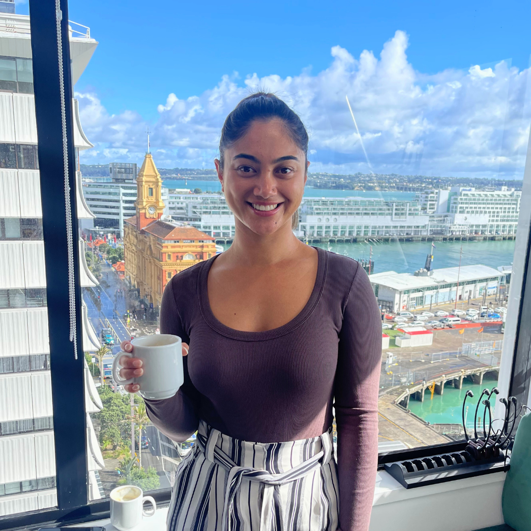 Woman holding a mug, smiling at the camera, standing by a window with a city and water view.