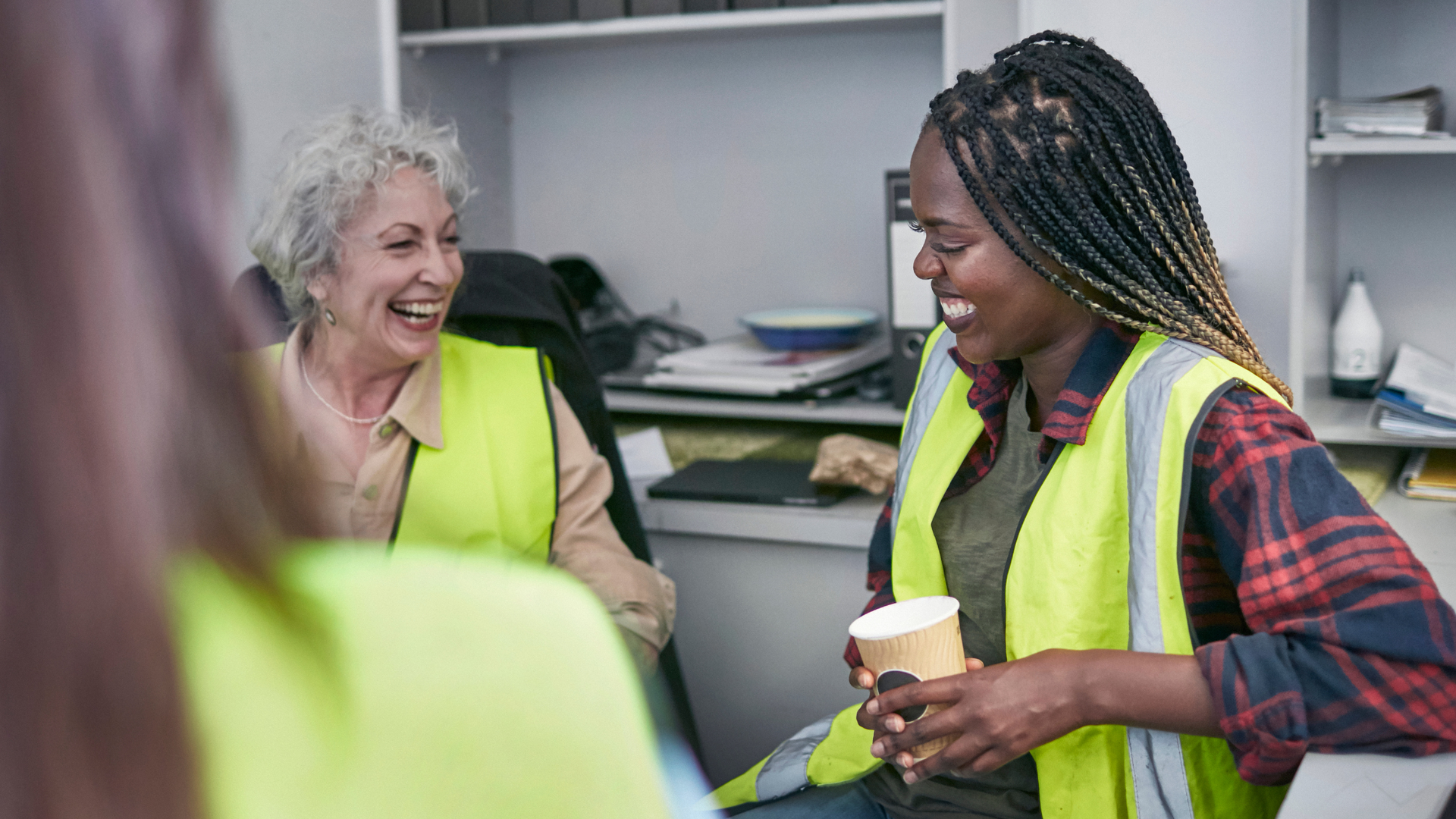 Two people in safety vests laugh, one holding a cup, in a well-lit office.