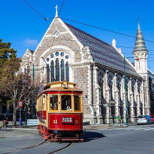 Red tram on tracks in front of a stone church with a steeple on a sunny day.