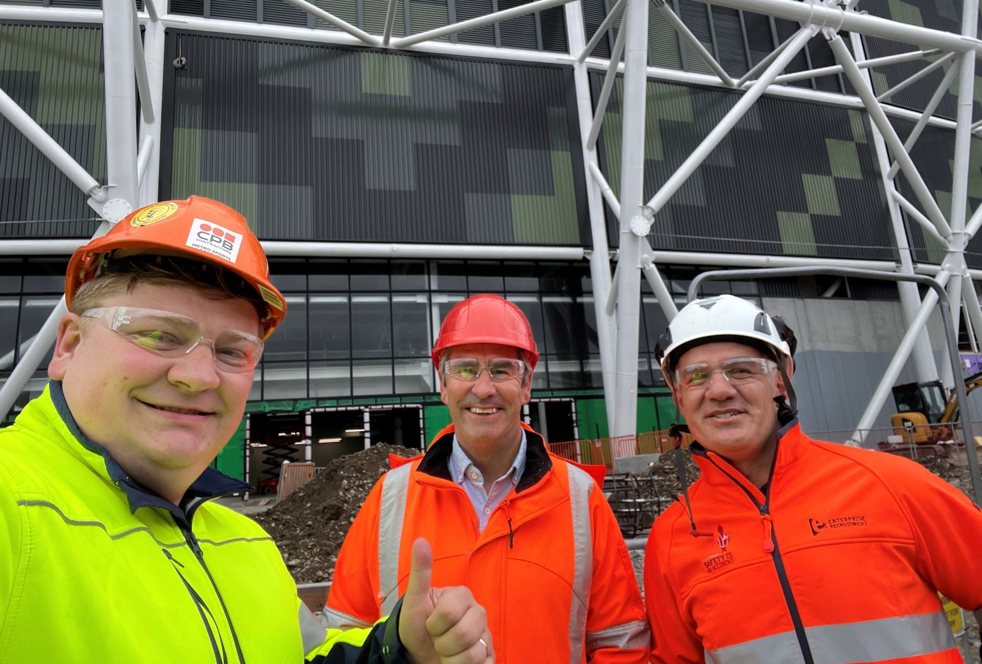 Three construction workers wearing hard hats and safety vests pose in front of a building.