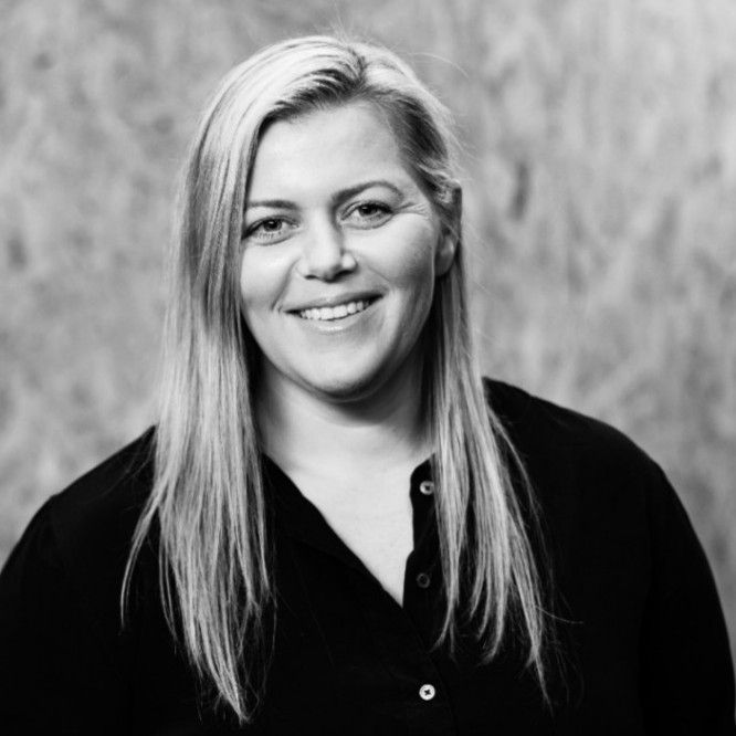 Woman with blonde hair smiling, wearing a black shirt, in front of a neutral background.
