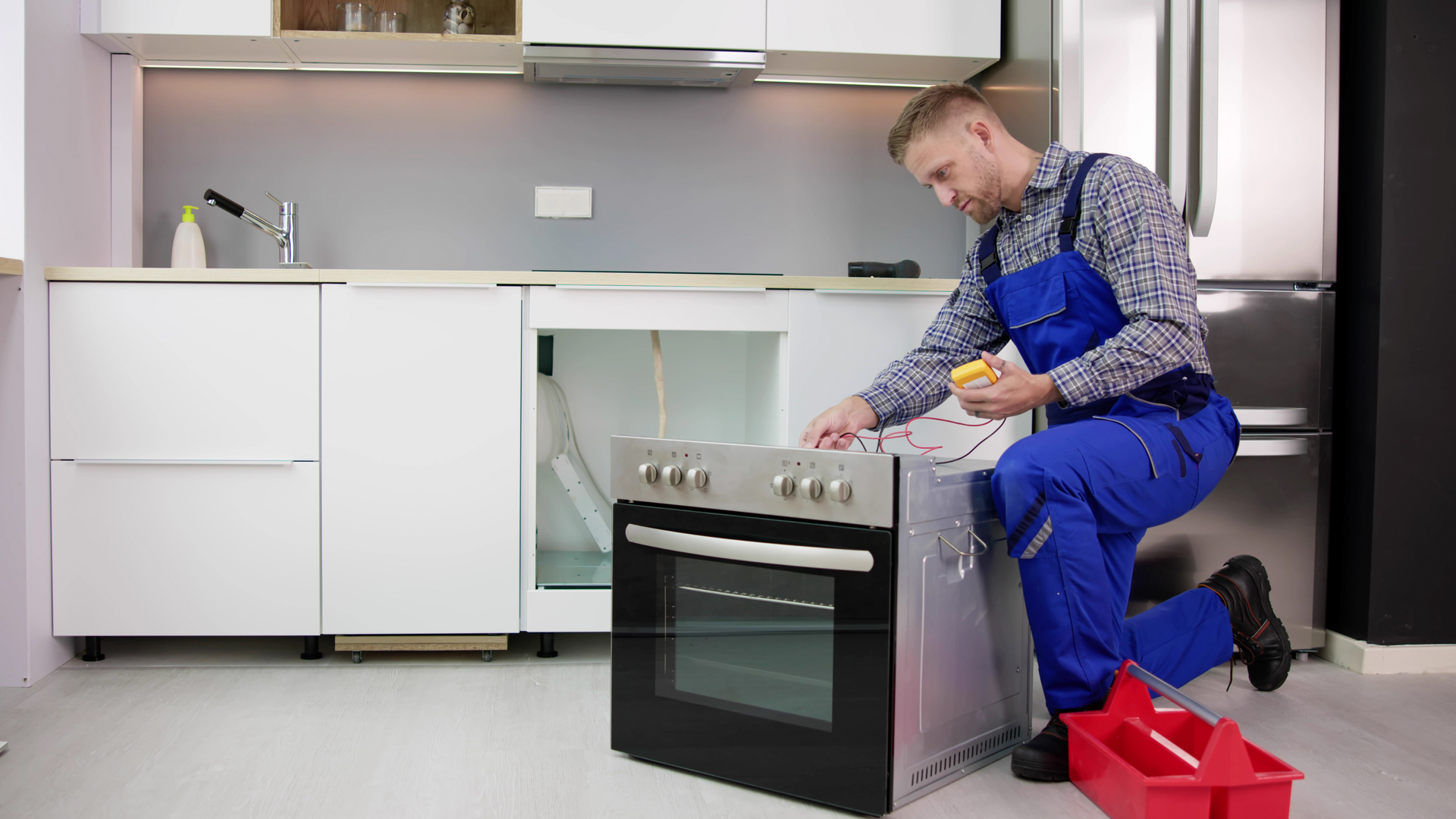 A repairman in blue overalls kneels, inspecting an oven in a kitchen. A toolbox sits nearby.