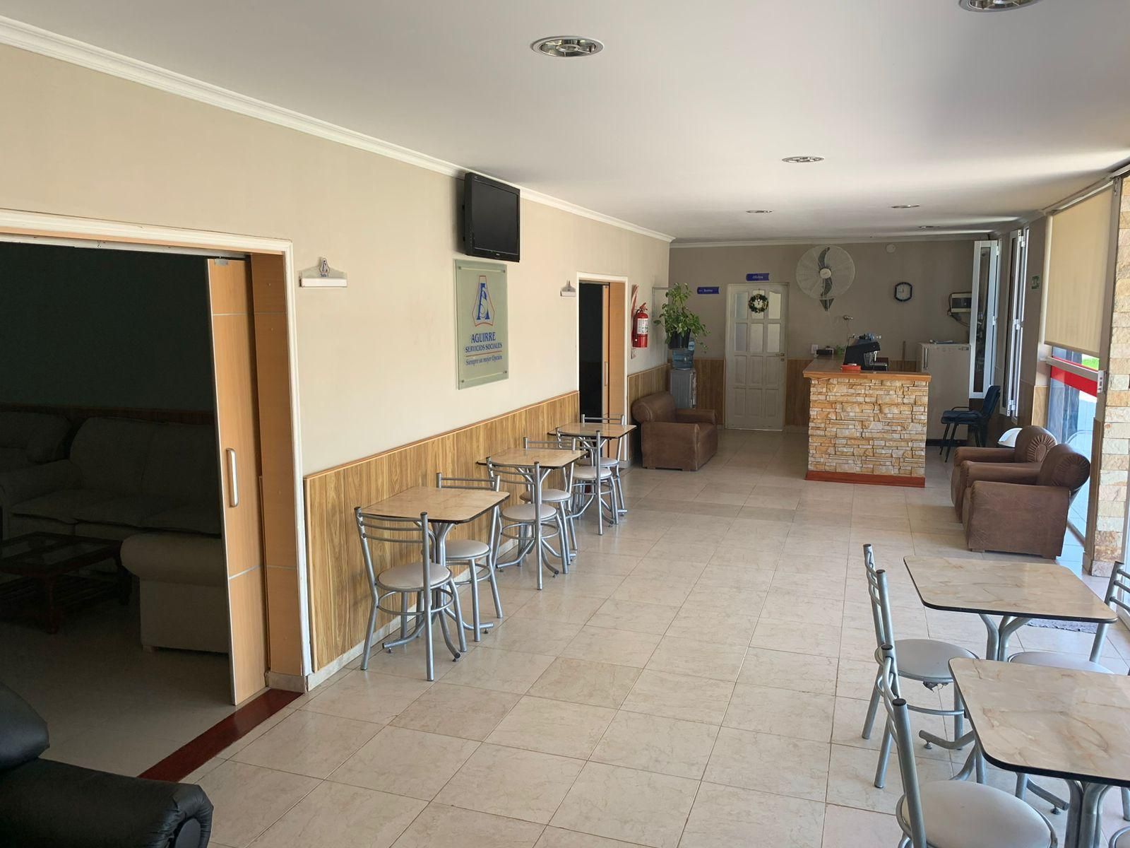 Zona de comedor de cafetería vacía, con suelo de baldosas, mesas y sillas de metal y una barra al fondo.