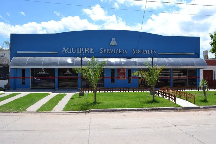 Fachada azul de Aguirre Services Socials con toldos, césped y una acera delante.