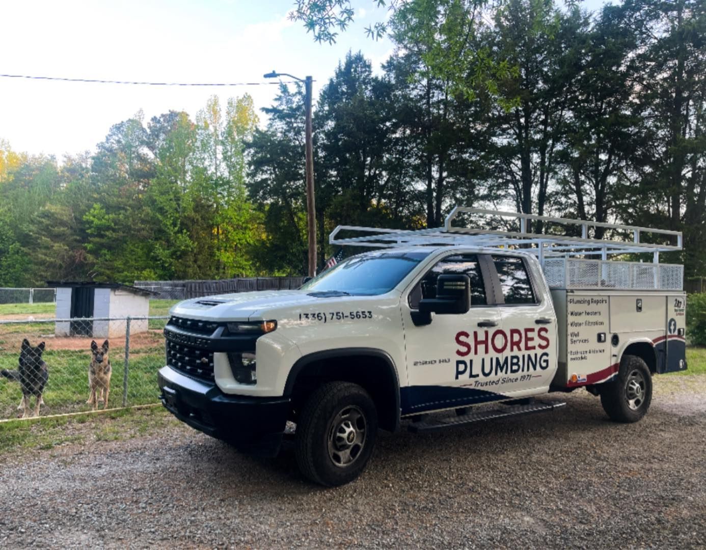 White Shores Plumbing service truck parked on a gravel driveway beside trees and a fence.