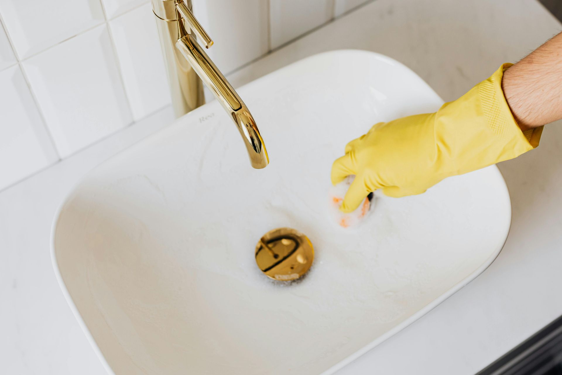 Gloved hand cleaning a white bathroom sink under a gold faucet
