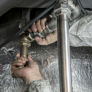Hands using a tool to tighten a metal pipe fitting under a sink, with water splashing nearby