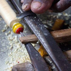 Hands carving a wooden tool handle with a knife and clamp close-up.