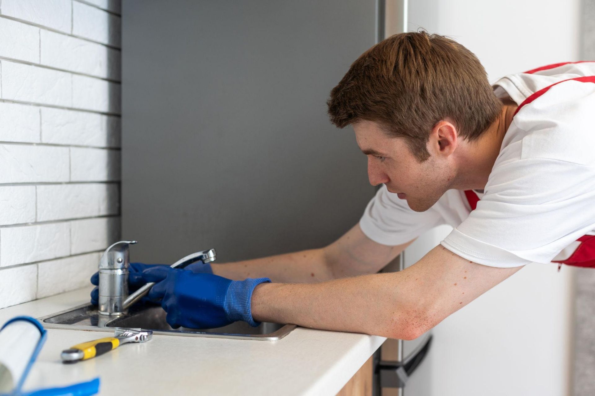 Plumber in blue gloves repairs a sink faucet in a bathroom with white tile walls