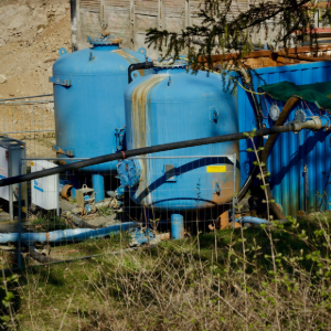 Blue industrial tanks and pipes beside a stone wall in a grassy outdoor utility area