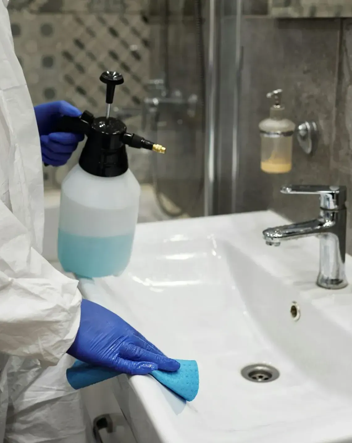 Person in blue gloves cleaning a white bathroom sink with a blue cloth and spray bottle nearby
