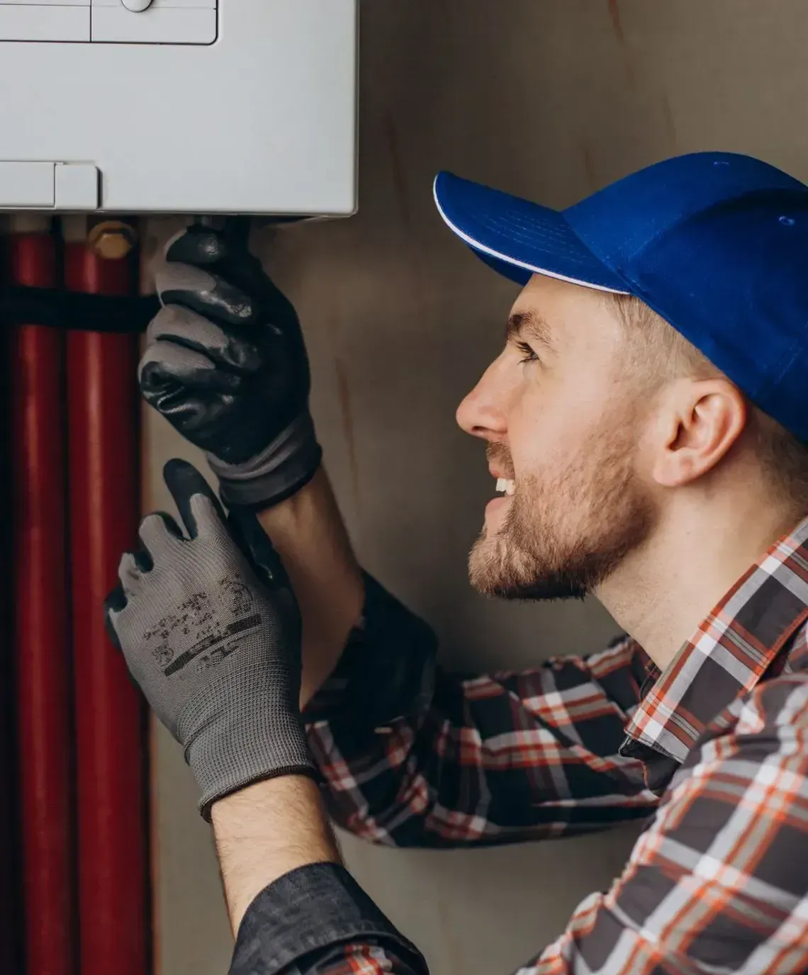 Technician in blue cap and gloves inspecting pipes and wiring inside a utility panel
