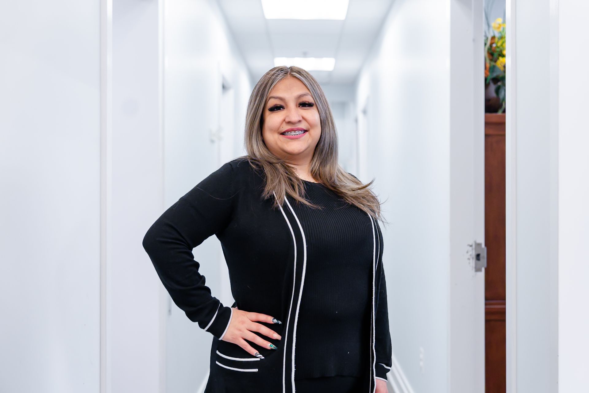 Woman in black sweater and skirt poses in a white hallway, smiling.