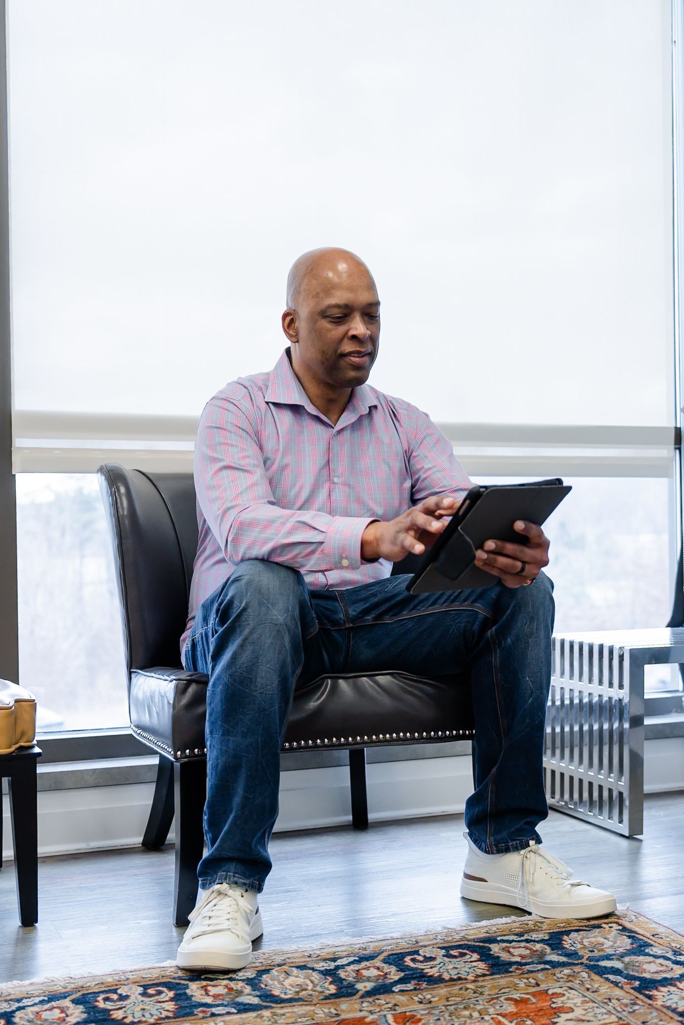 Man in a button-up shirt and jeans sits in a chair, using a tablet. White sneakers, window with blinds.