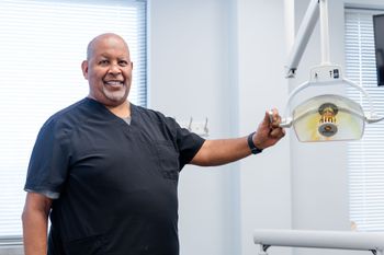 Dentist in black scrubs smiles, adjusting dental lamp in a bright office.
