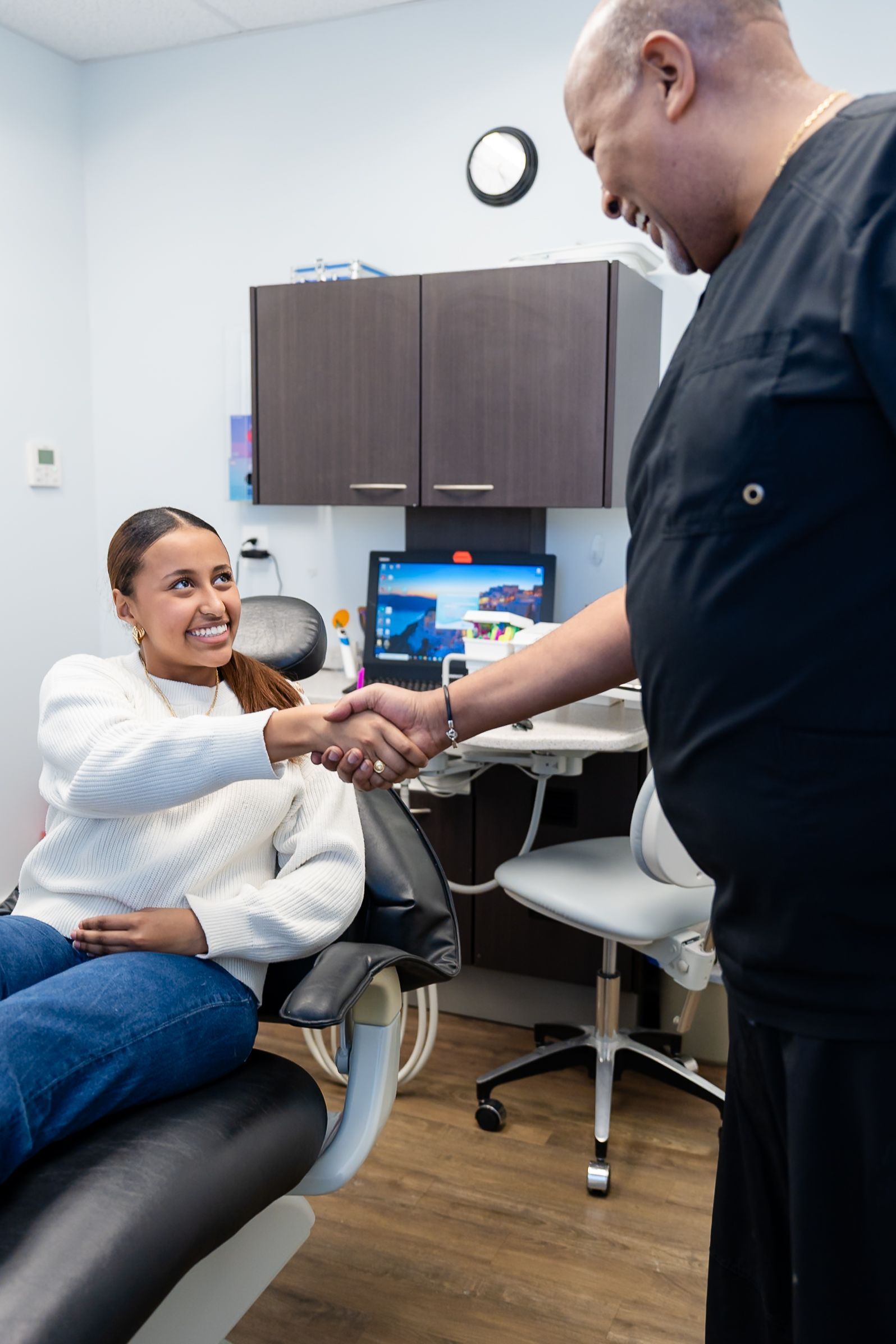 A smiling person in dental chair shakes hands with a person in scrubs in an office.