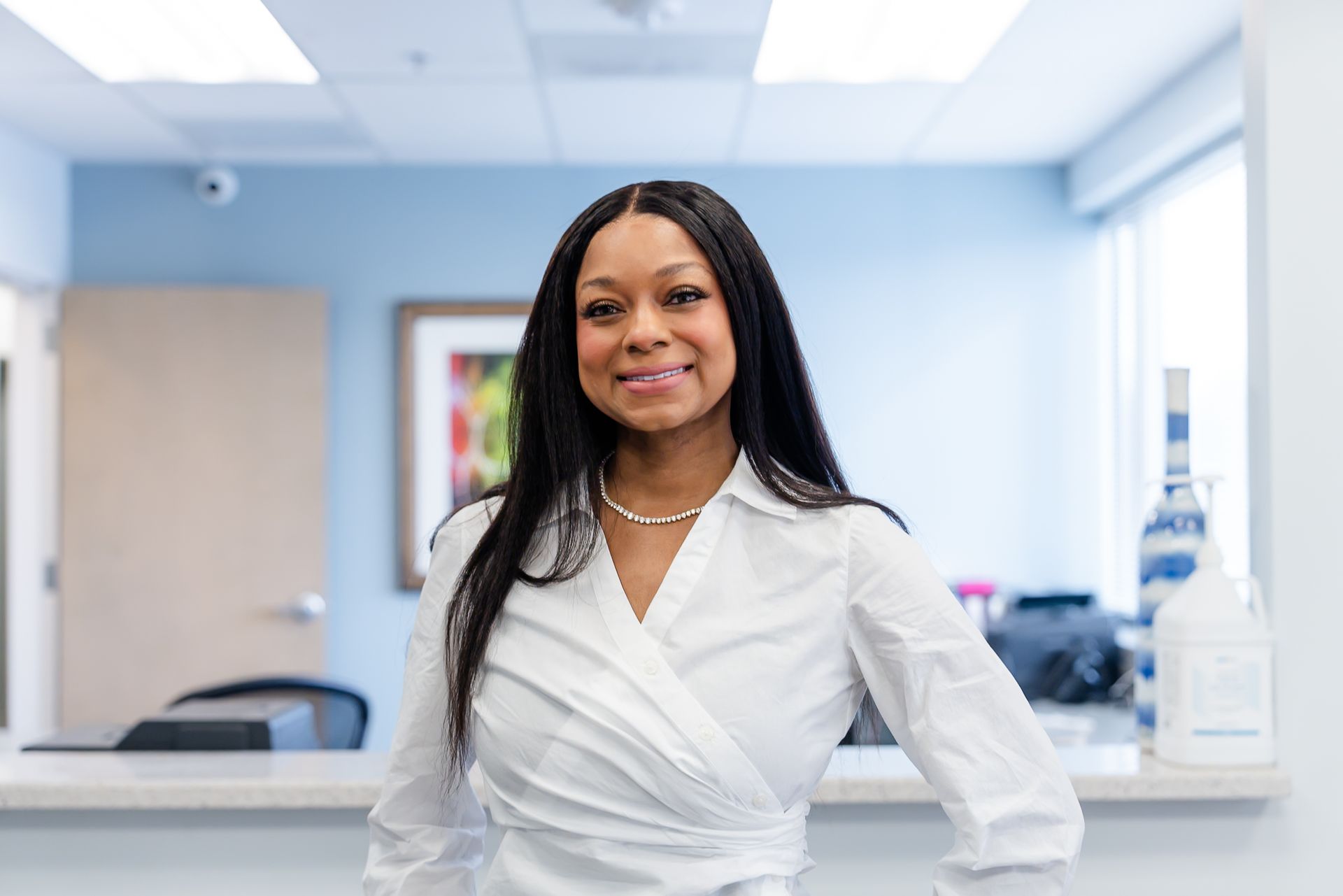 Woman in white blouse smiles at reception desk. Office setting, light blue walls, and art.